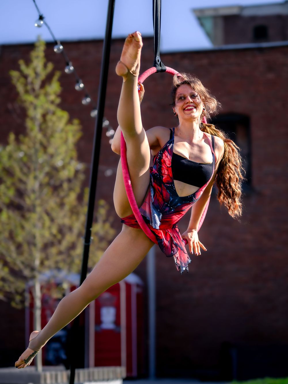 Aerial acrobat performs graceful splits on a lyra hoop outdoors, wearing a red and black costume against a brick building.