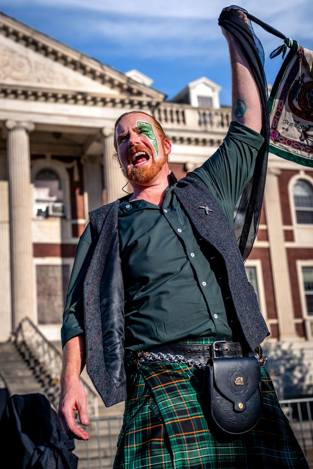 Man in tartan kilt and green face paint raises flag, celebrating St. Patrick's Day outdoors near historic building.
