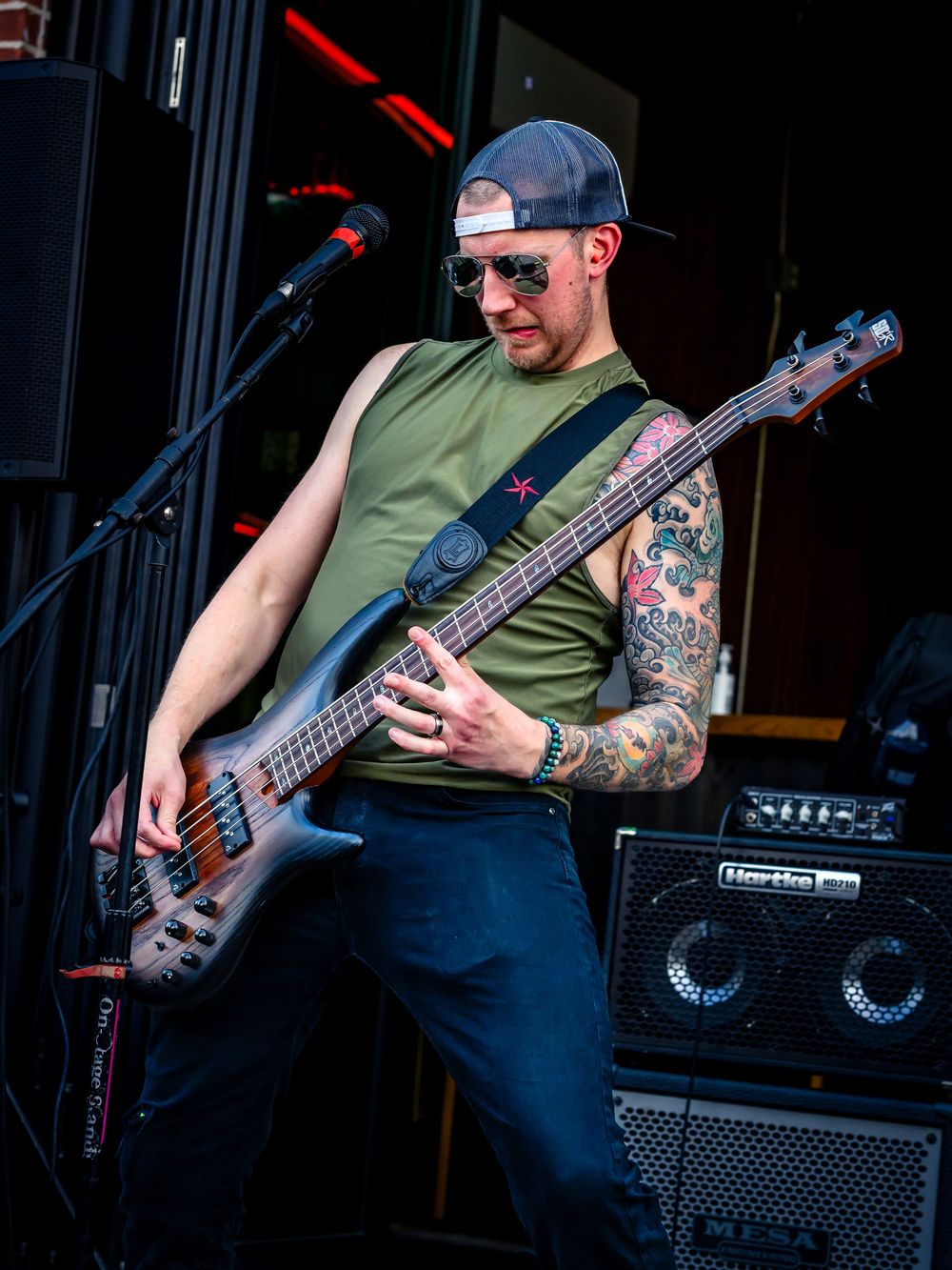 Tattooed bassist performs on stage wearing a green sleeveless shirt and cap, playing a dark electric bass guitar.