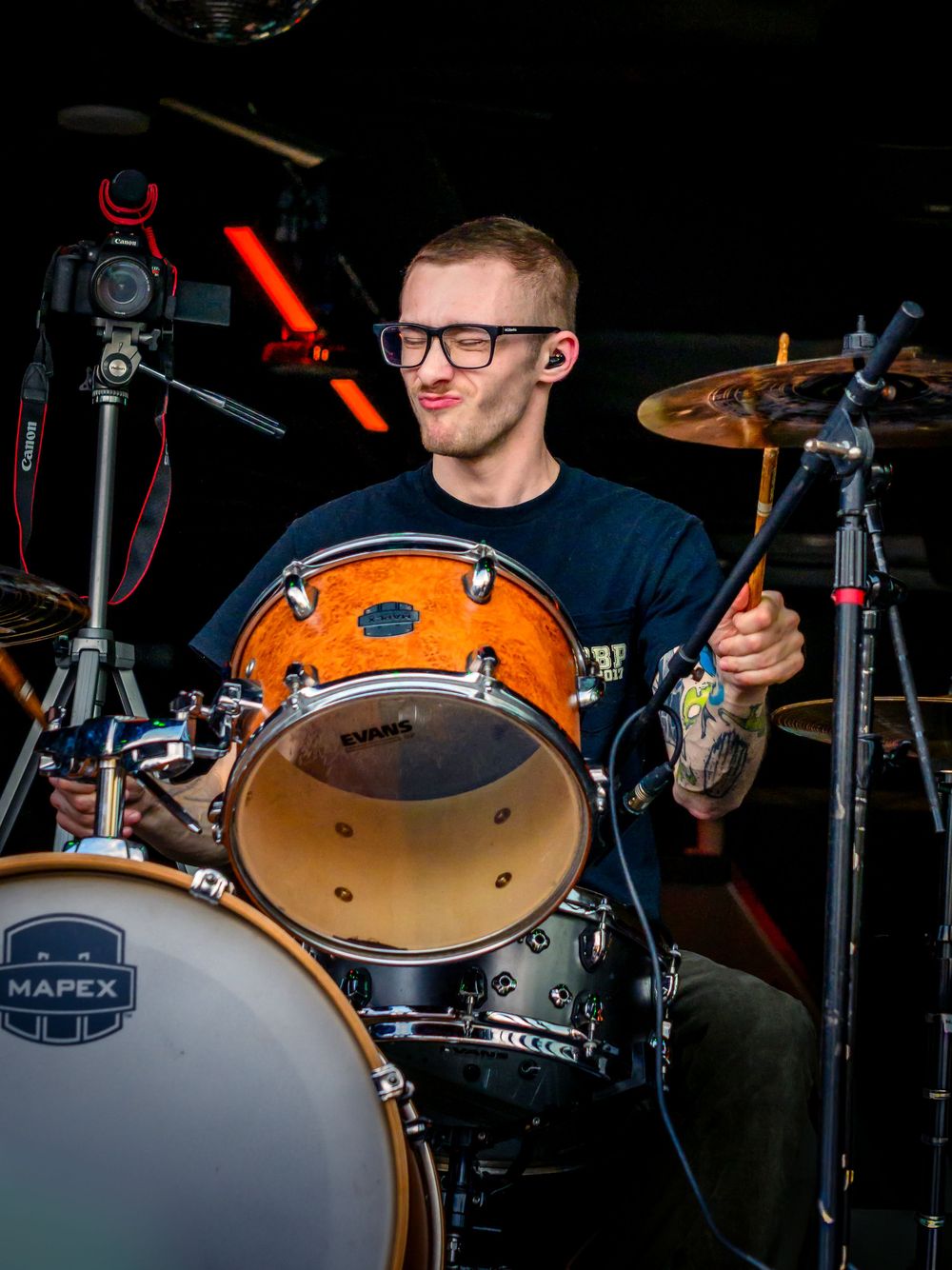 Young male drummer with glasses playing Mapex drums on stage during a live performance.