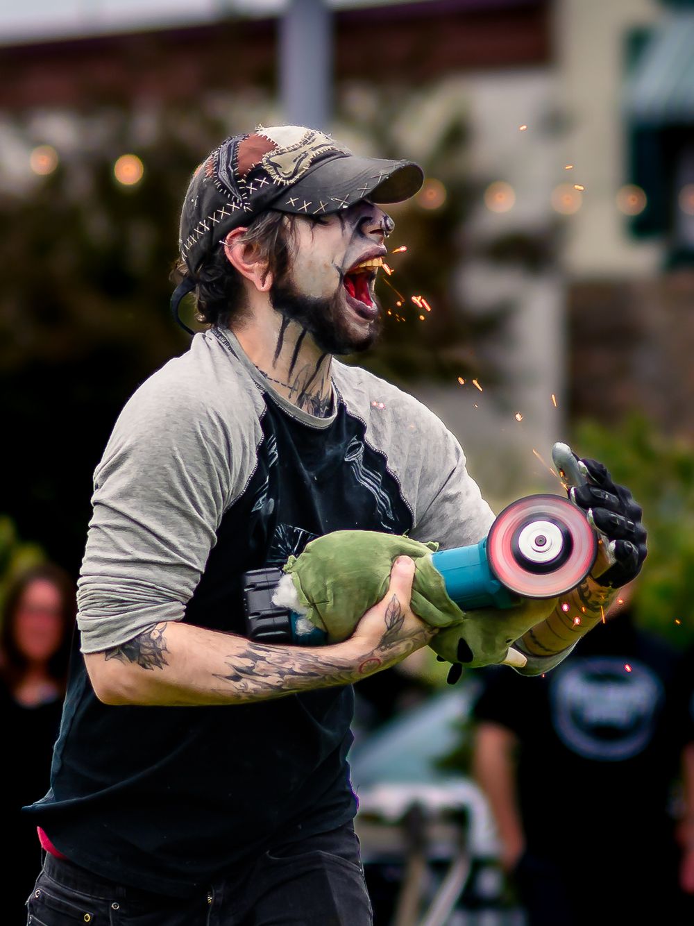 Tattooed man with cap reacts dramatically while holding angle grinder with sparks flying outdoors.