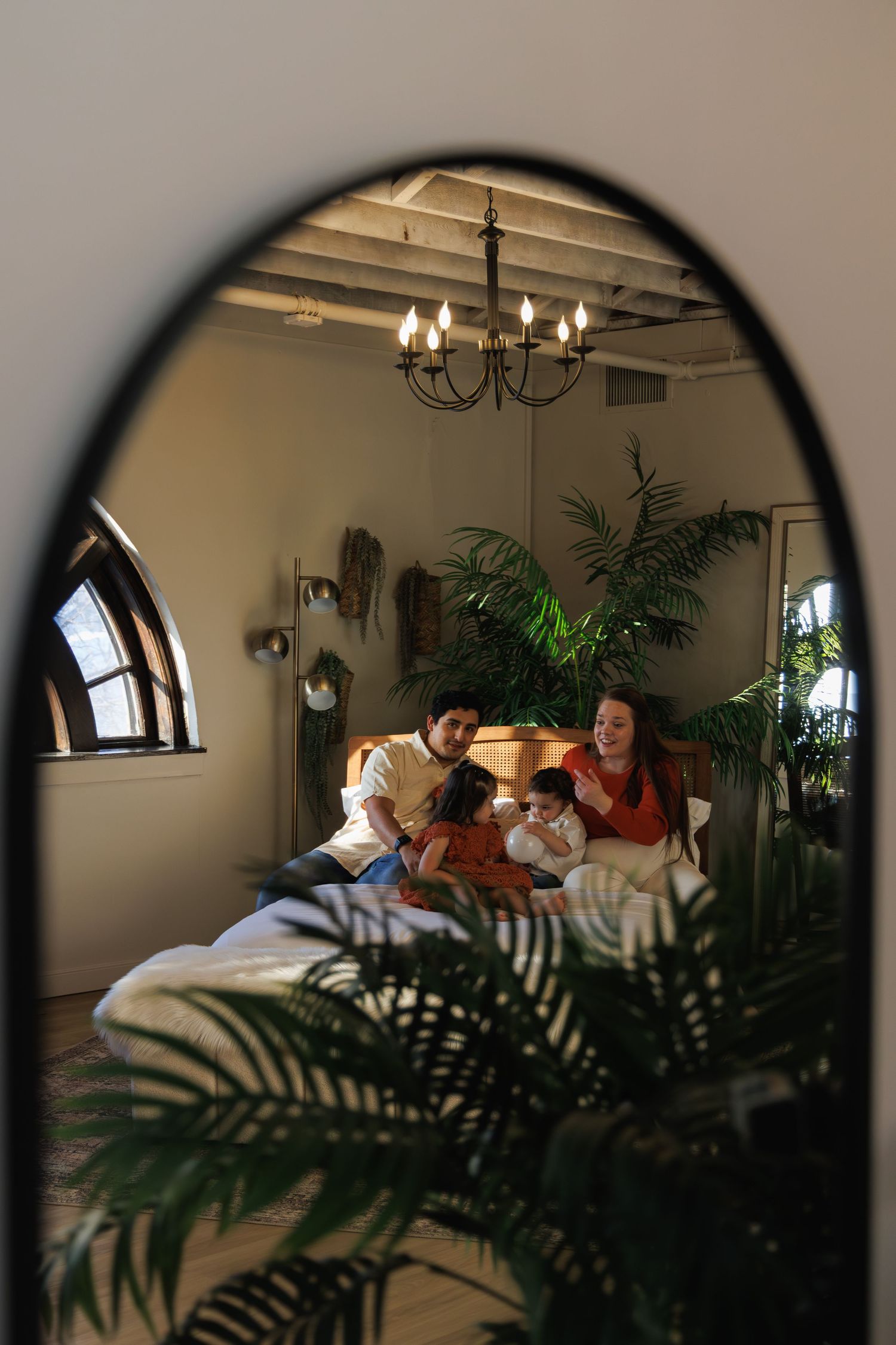 A cozy bedroom scene viewed through an arched mirror with houseplants and chandelier.