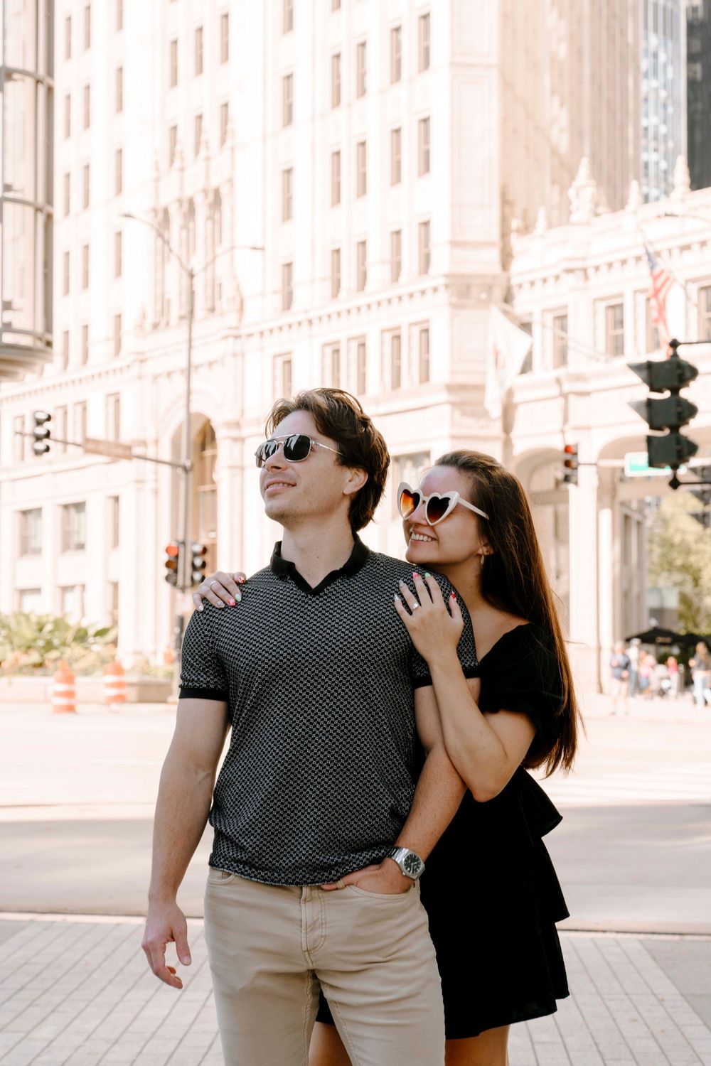 Stylish couple in sunglasses share a romantic moment on a city street with tall buildings in the background.