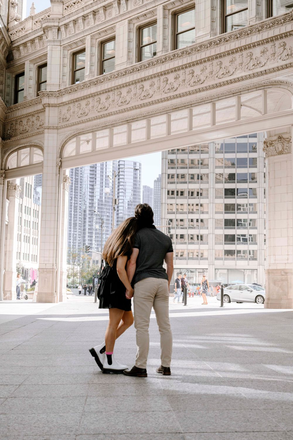 A couple embraces while standing beneath an ornate archway of a historic Chicago building with skyscrapers in view.