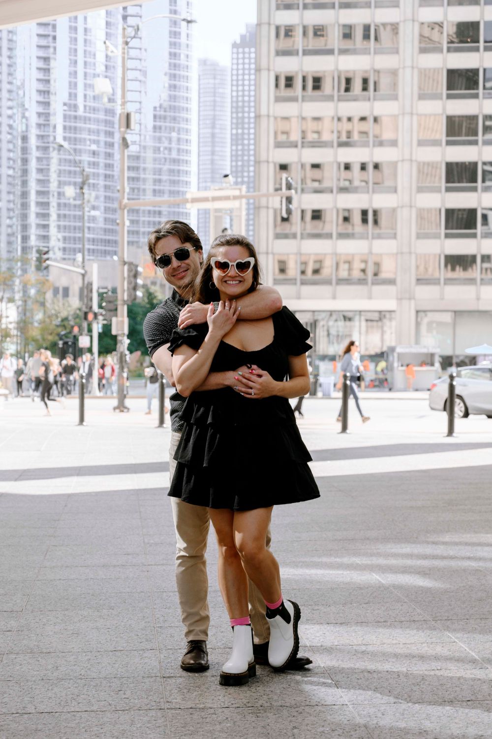 Happy couple wearing sunglasses embrace on an urban street corner with skyscrapers in the background.