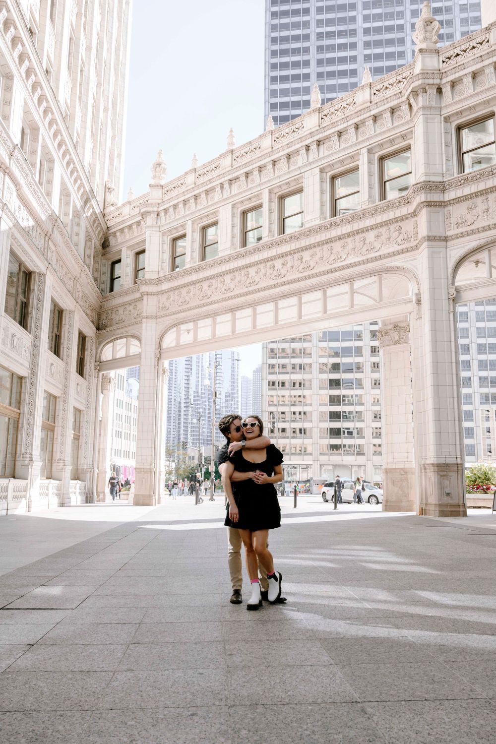 A couple embraces under an ornate architectural archway in downtown Chicago with modern skyscrapers visible in the background.