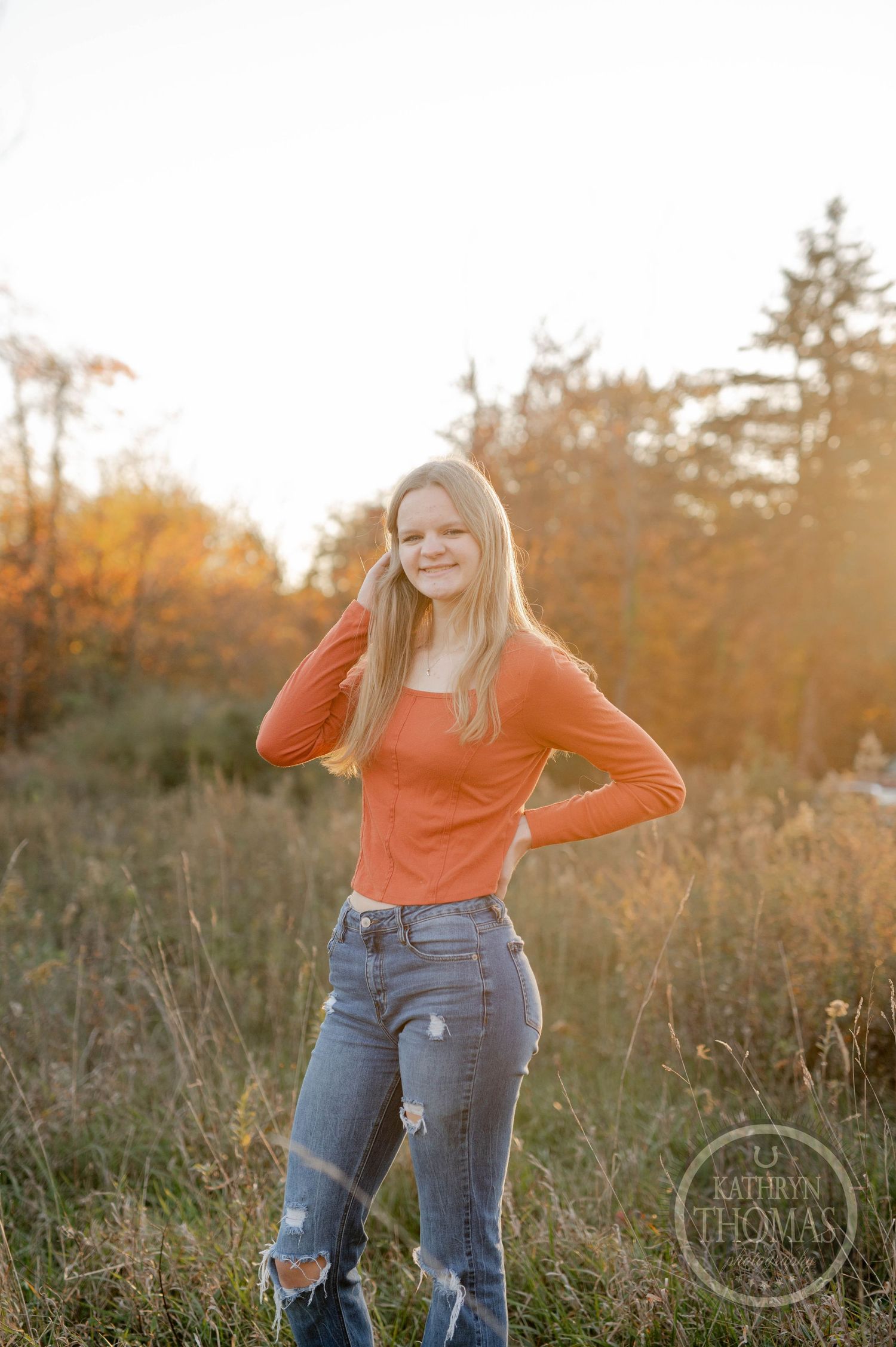 Person in coral sweater and distressed jeans poses confidently in autumn field at sunset.