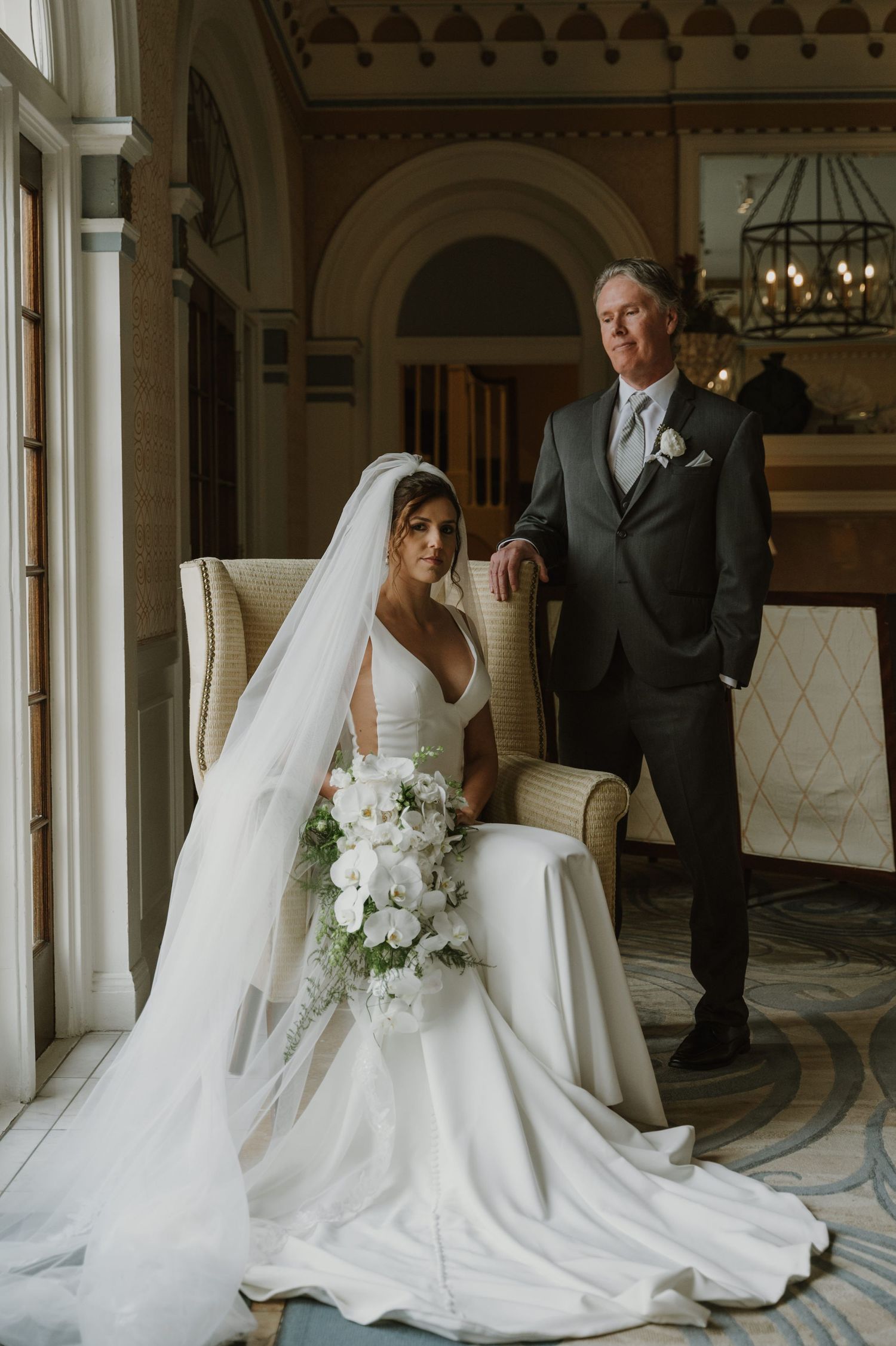 Elegant wedding portrait in an ornate venue featuring a bride in a flowing white gown seated beside a suited companion.