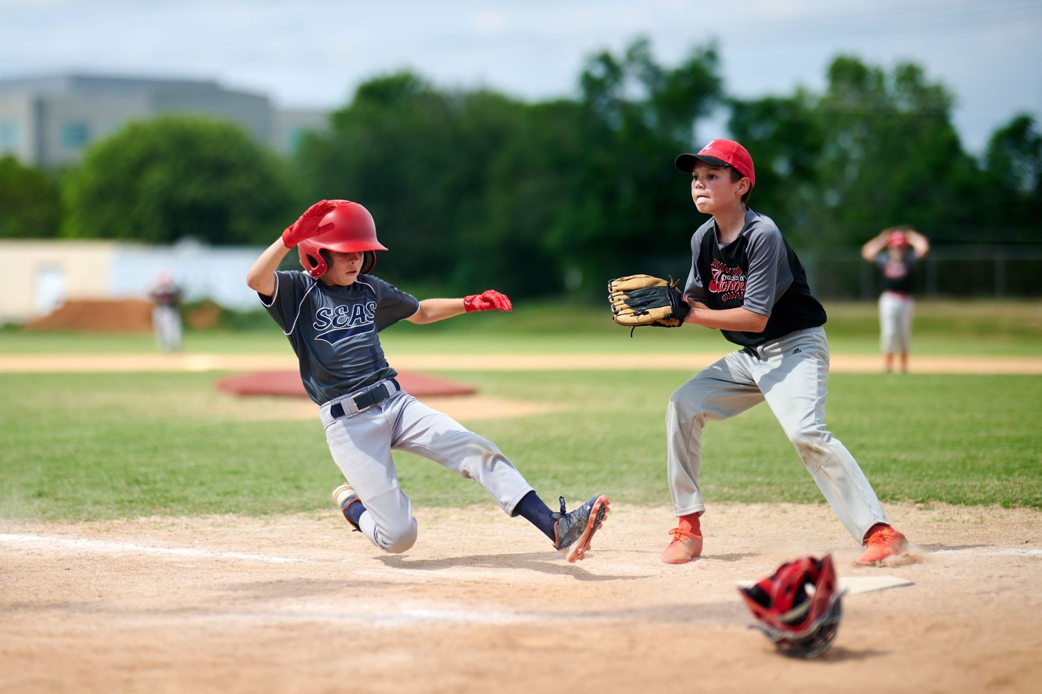 Two youth baseball players in action during a game, with one sliding into base while another fields the ball.