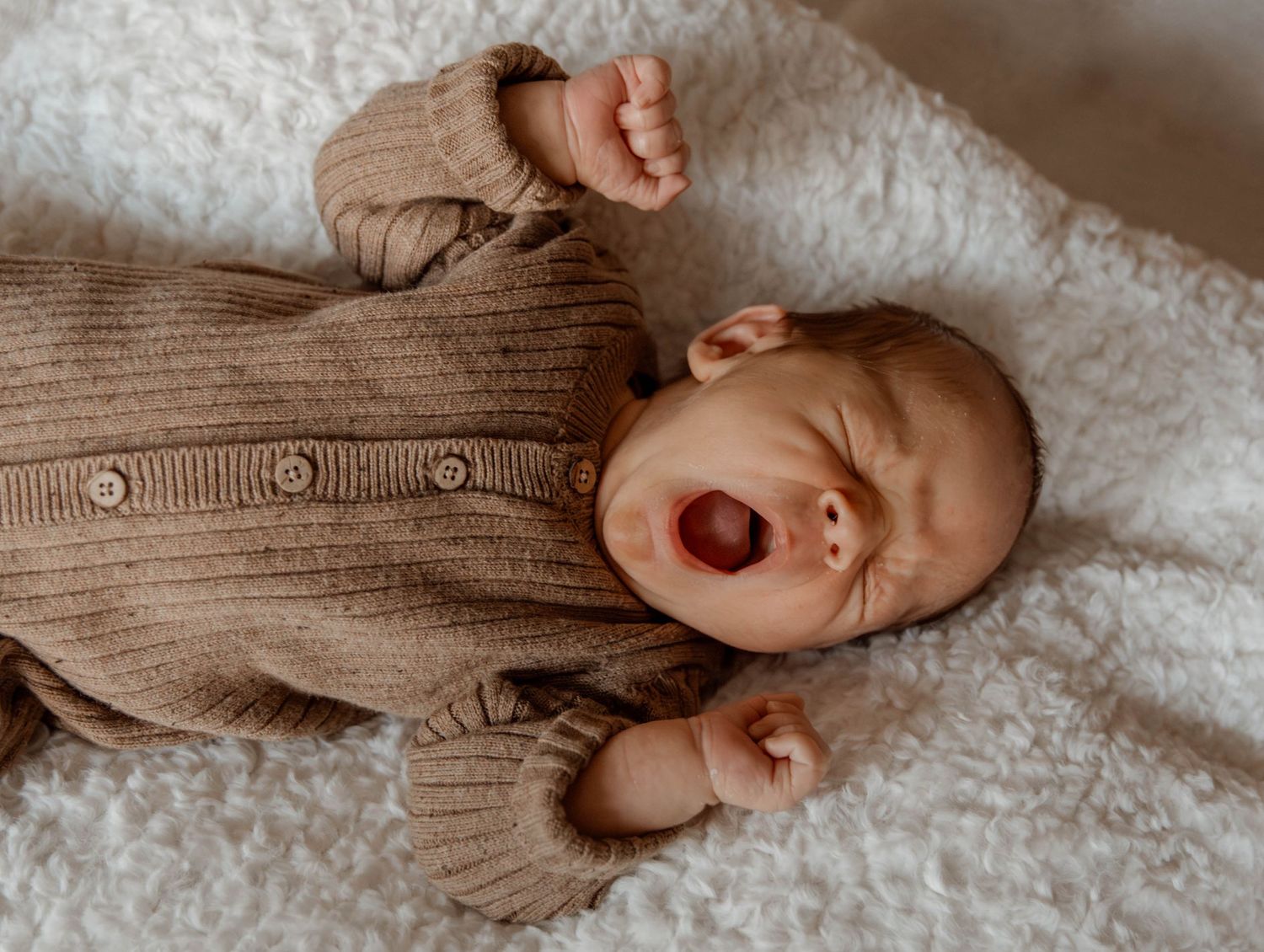 Yawning baby in beige sweater lying on white textured blanket.