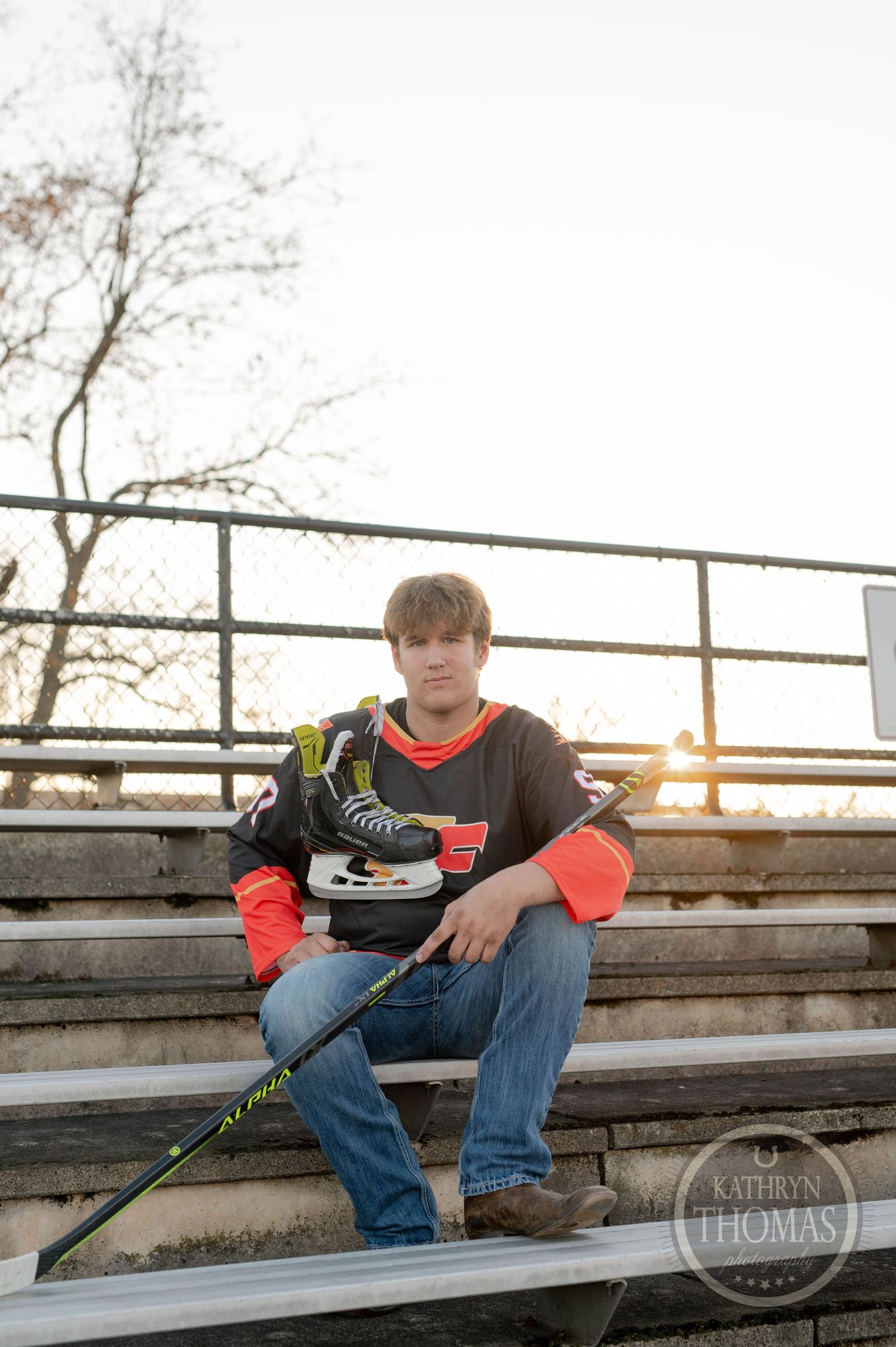 High school athlete poses on metal bleachers with hockey gear during sunset.