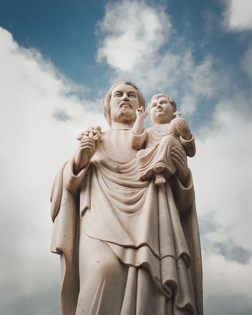 Classical stone statues draped in flowing robes against a cloudy blue sky.