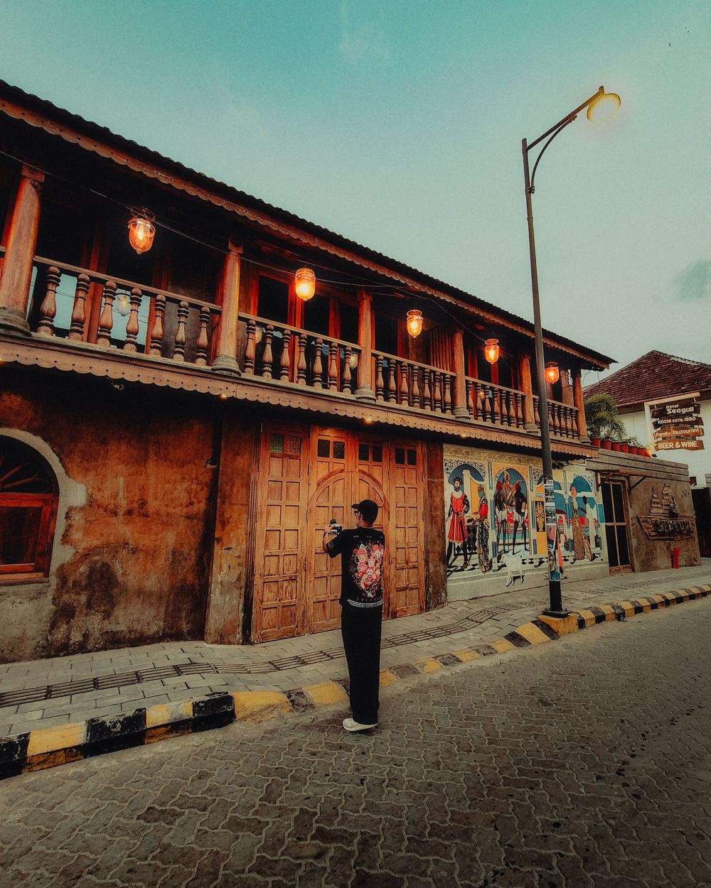 Person standing on a cobblestone street beside a vintage two-story building with lanterns and colorful murals at dusk.
