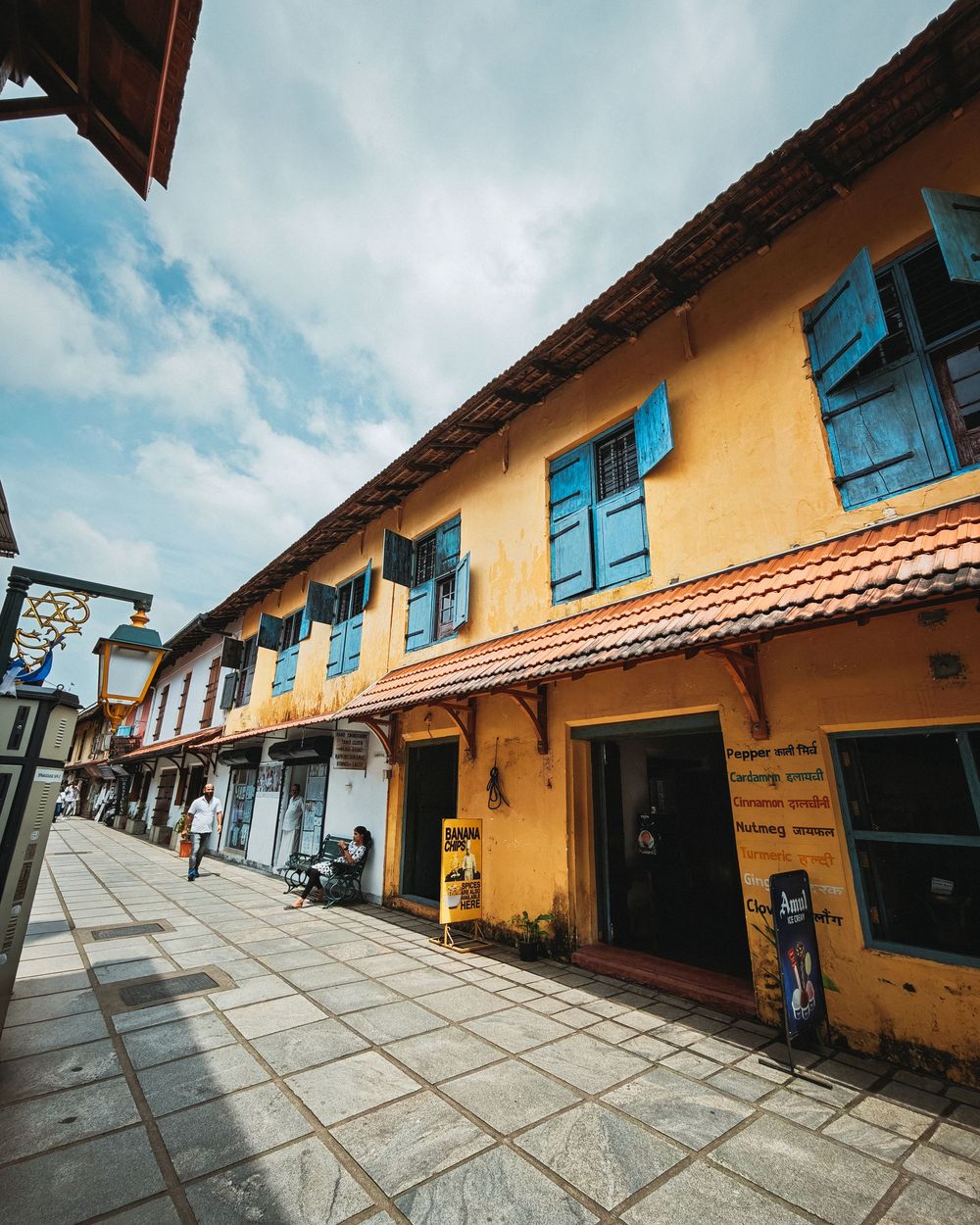 Panoramic view of yellow colonial buildings with blue shutters and terracotta roof tiles along a paved courtyard walkway.