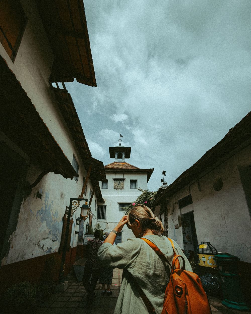 Person with backpack walks through narrow alley towards white tower under moody sky.