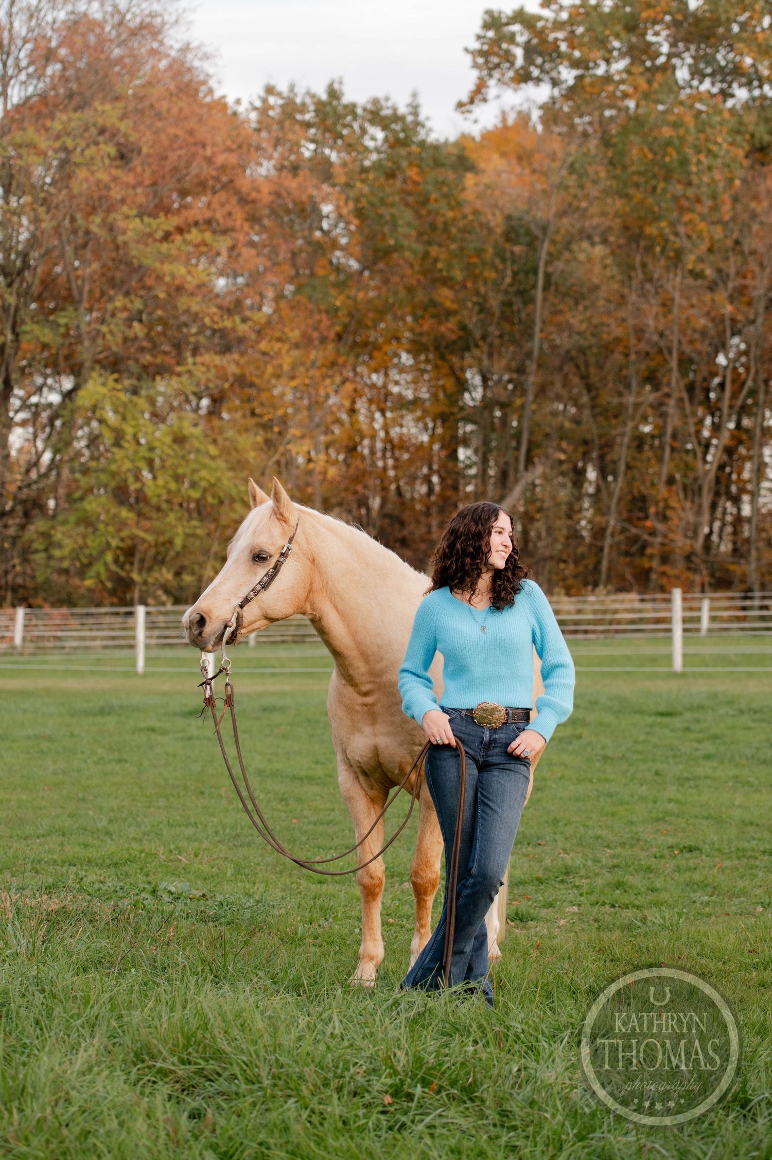 A palomino horse stands in profile next to someone in a blue sweater in an autumn field.