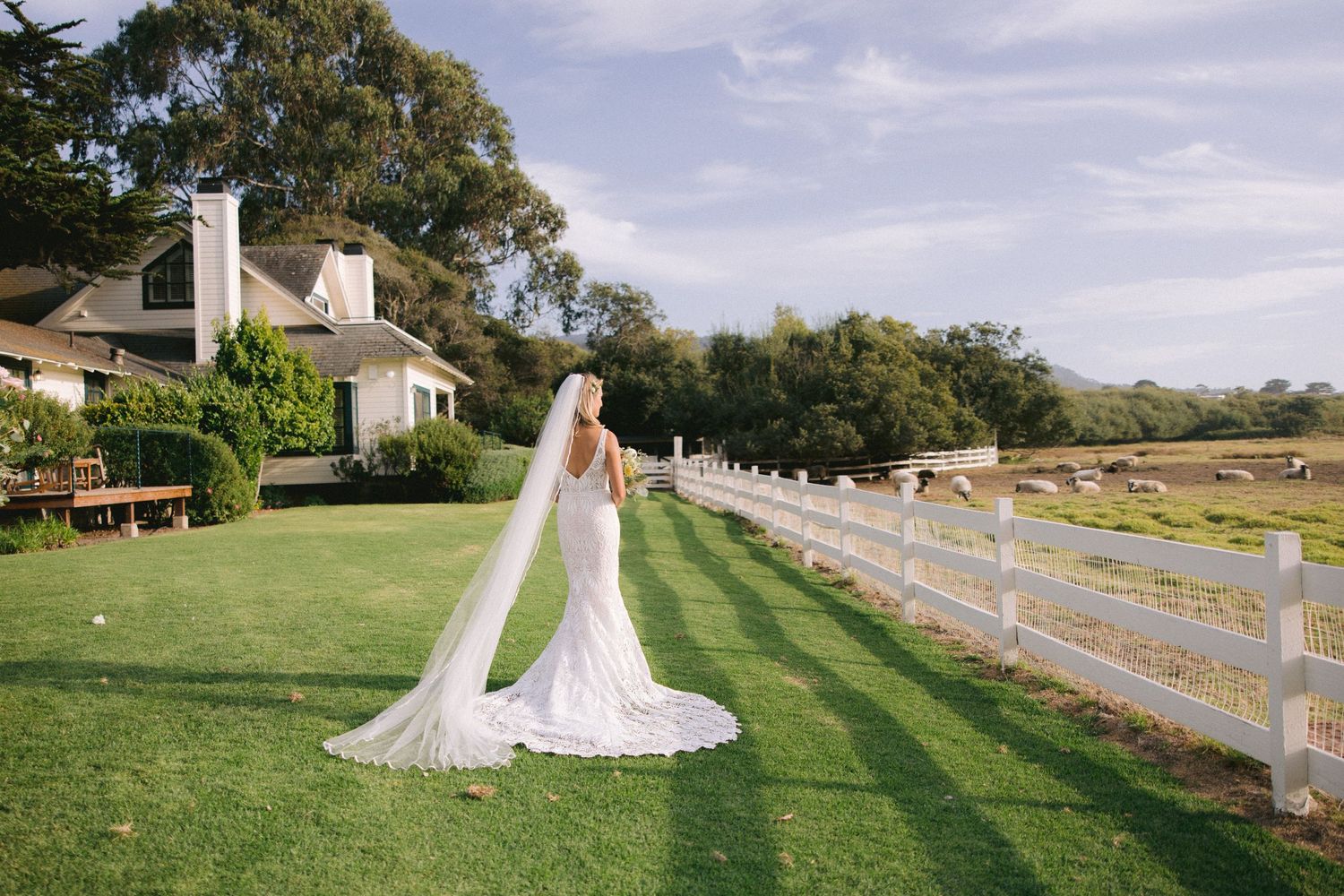 A flowing white wedding gown stands out against green lawn and white fence at a rustic countryside venue.