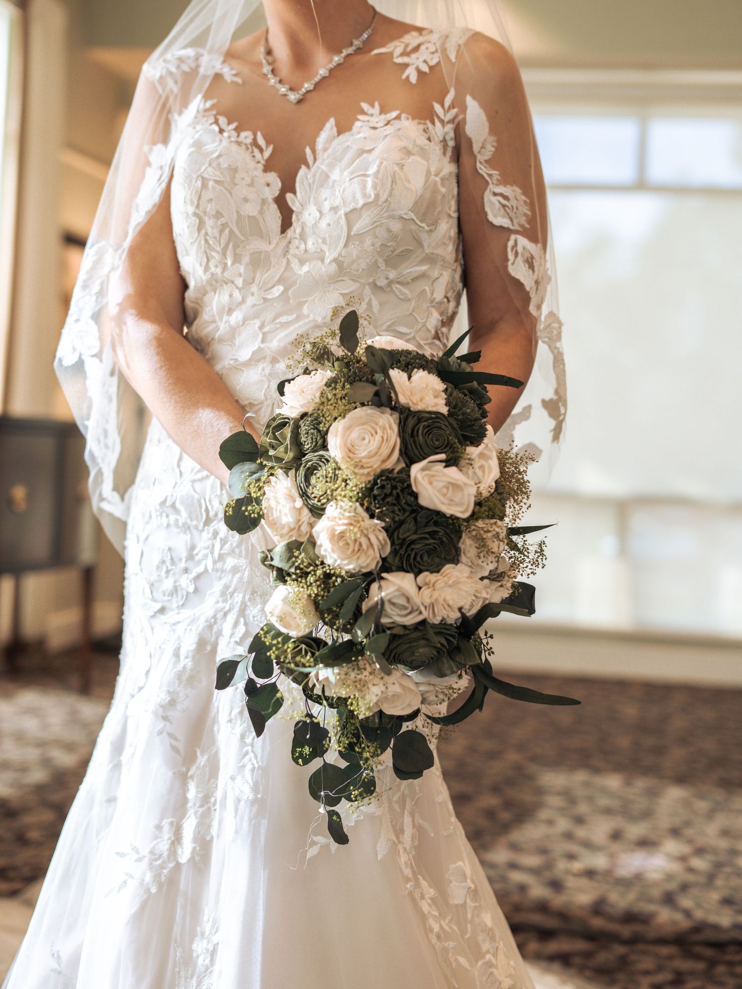 Close-up view of a bridal bouquet with white roses and eucalyptus against a lace wedding gown with sheer details.