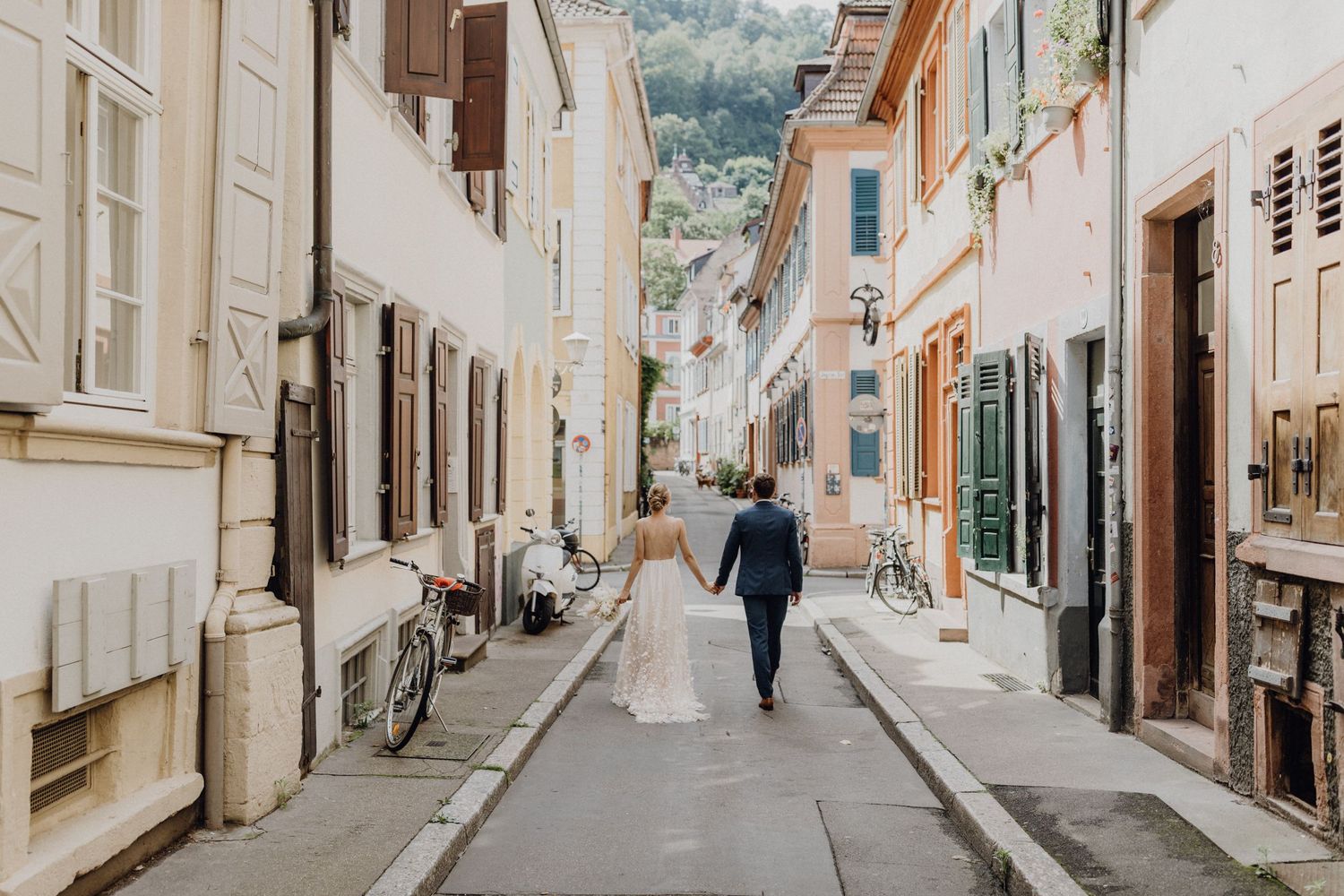 Brautpaar, das Hand in Hand durch eine Altstadtgasse in Heidelberg läuft. Die Sonne scheint, es ist Sommer.