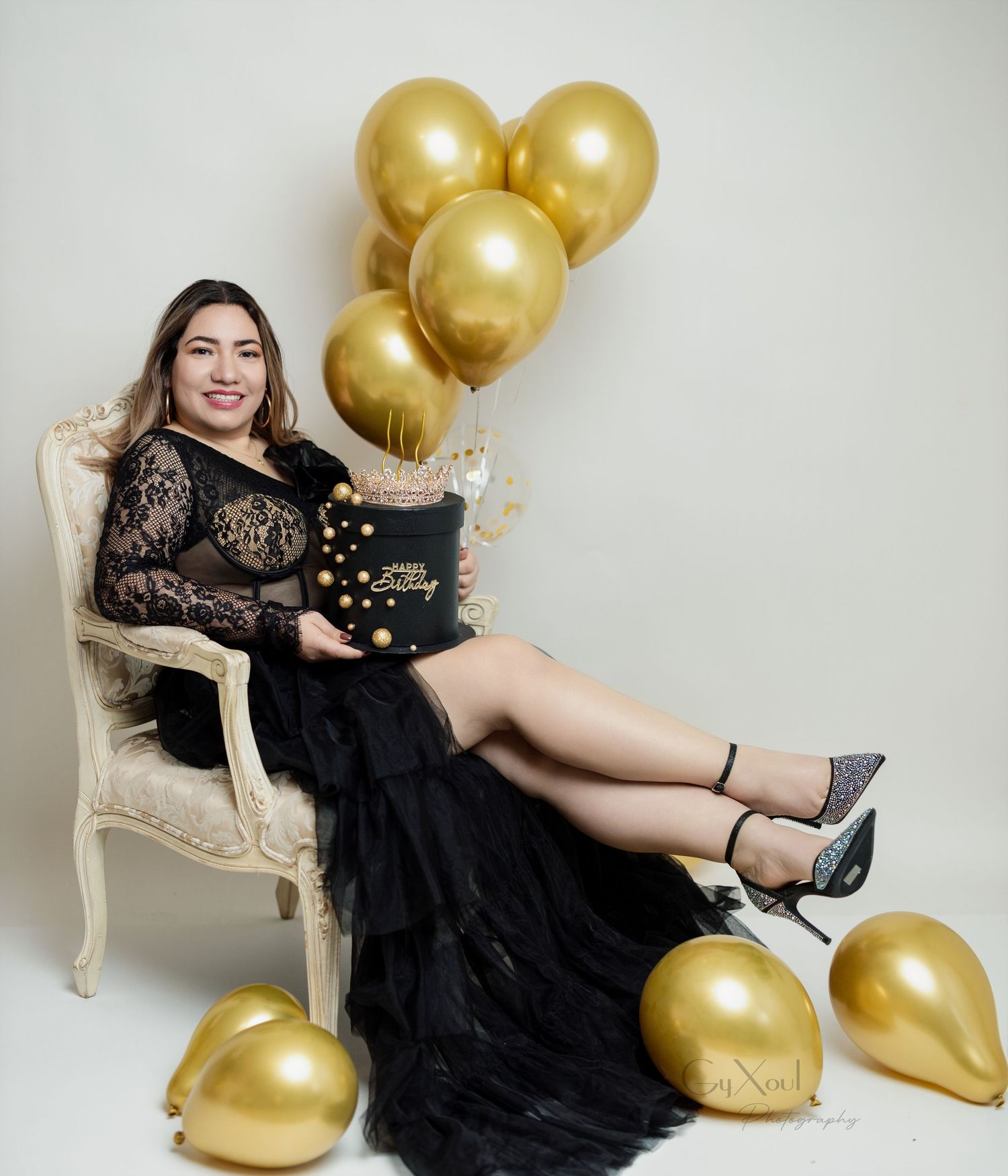 Elegant celebration setup with black lace dress, vintage chair, gold balloons and decorative cake.