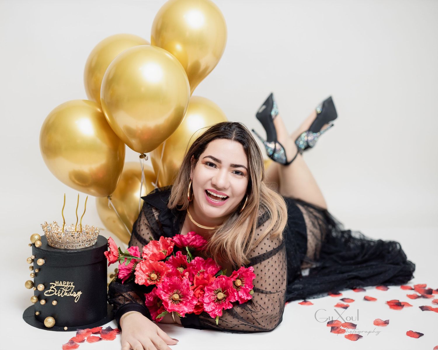 Birthday photoshoot with gold balloons, pink flowers and black lace outfit on white background.