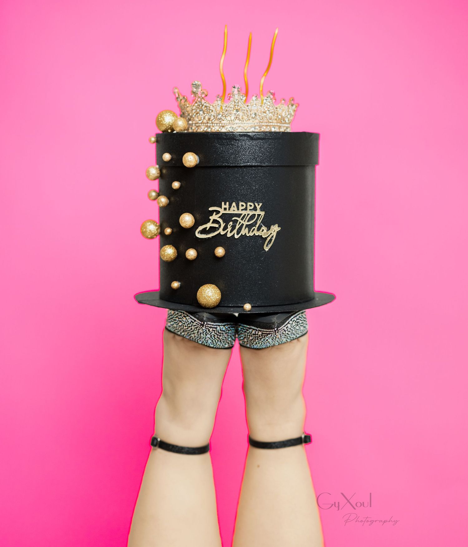 Black and gold birthday cake held up by legs wearing strappy heels against pink background.