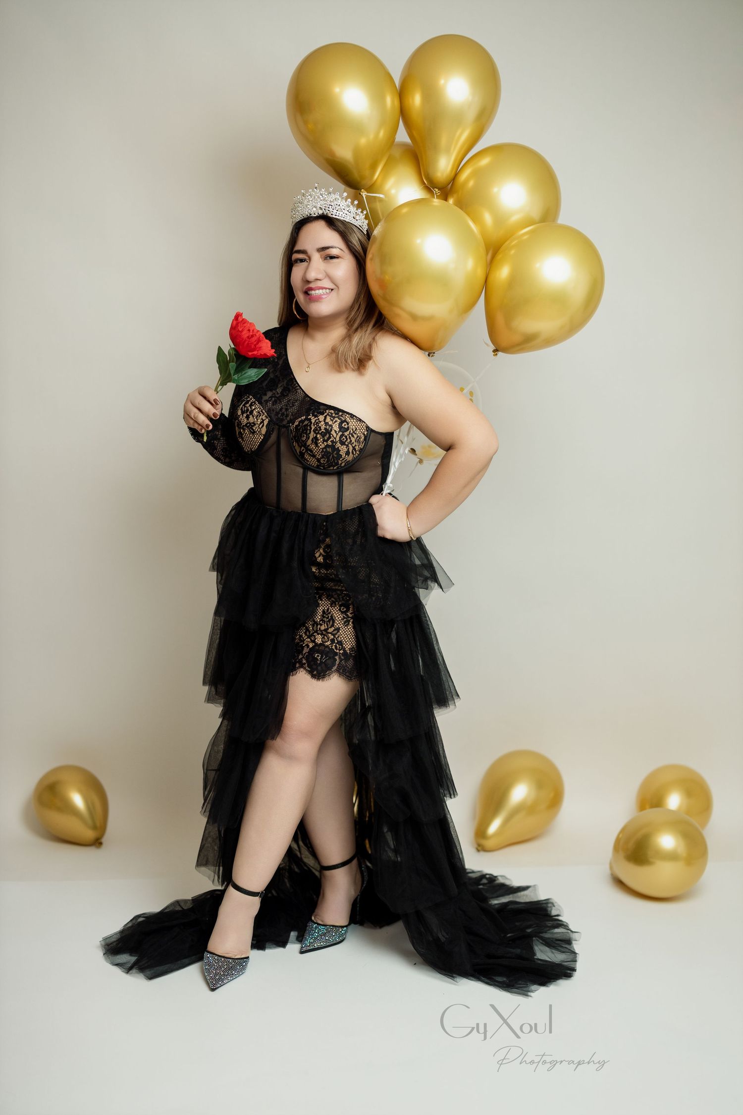 Celebratory portrait with black tulle dress, red rose and gold balloons against neutral background.