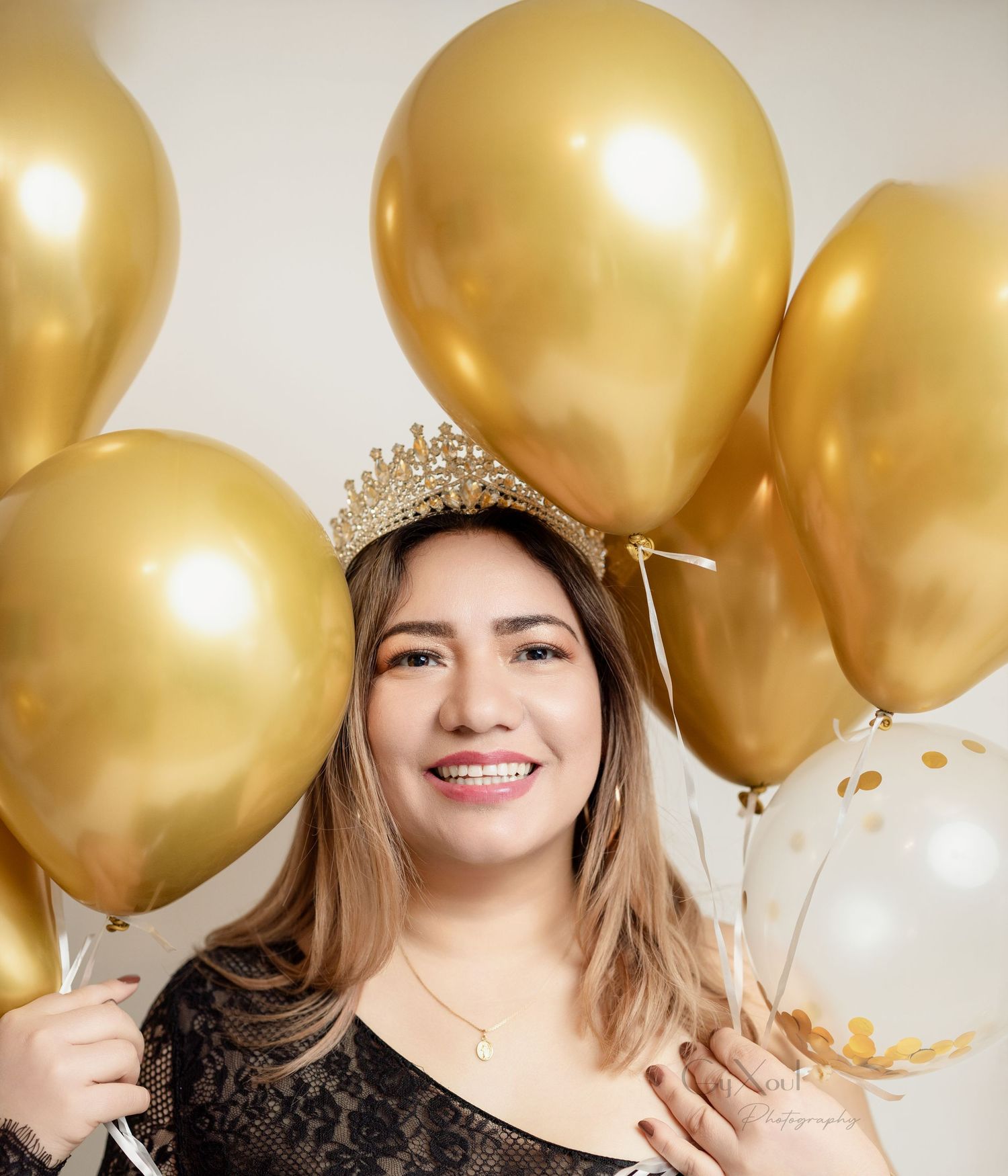 A festive portrait with gold balloons and a sparkly crown against a neutral background.