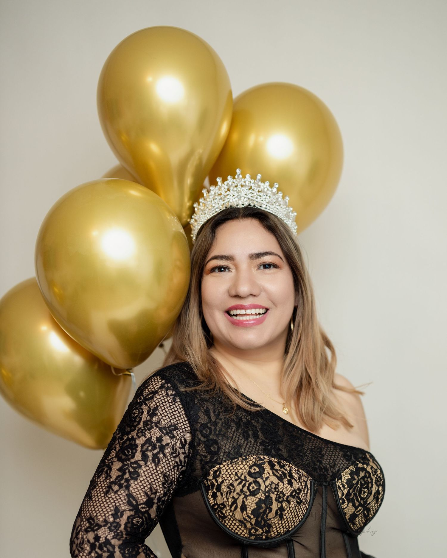 Glamorous portrait with crystal crown, black lace top and golden balloons in background.