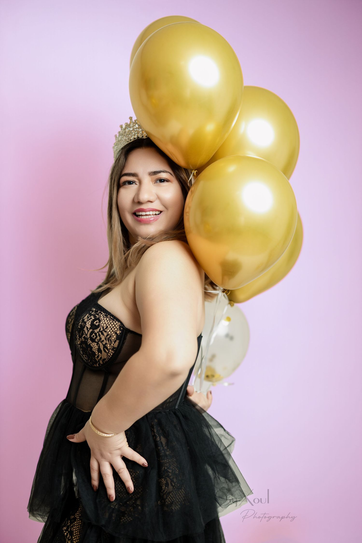 A celebratory portrait with gold balloons against a pink background in a black lace dress.