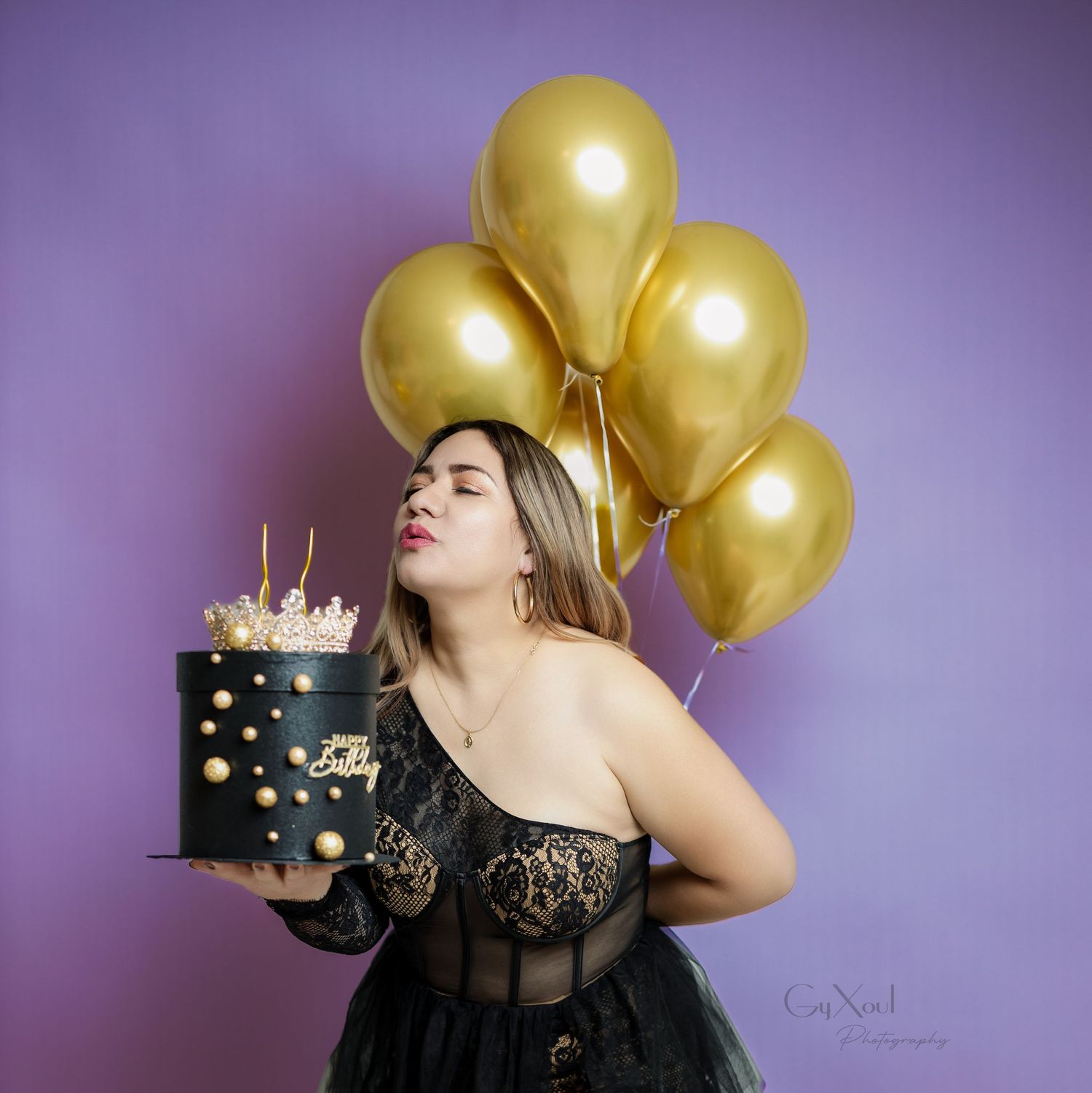 A birthday celebration scene with gold balloons and a black decorated cake against purple backdrop.