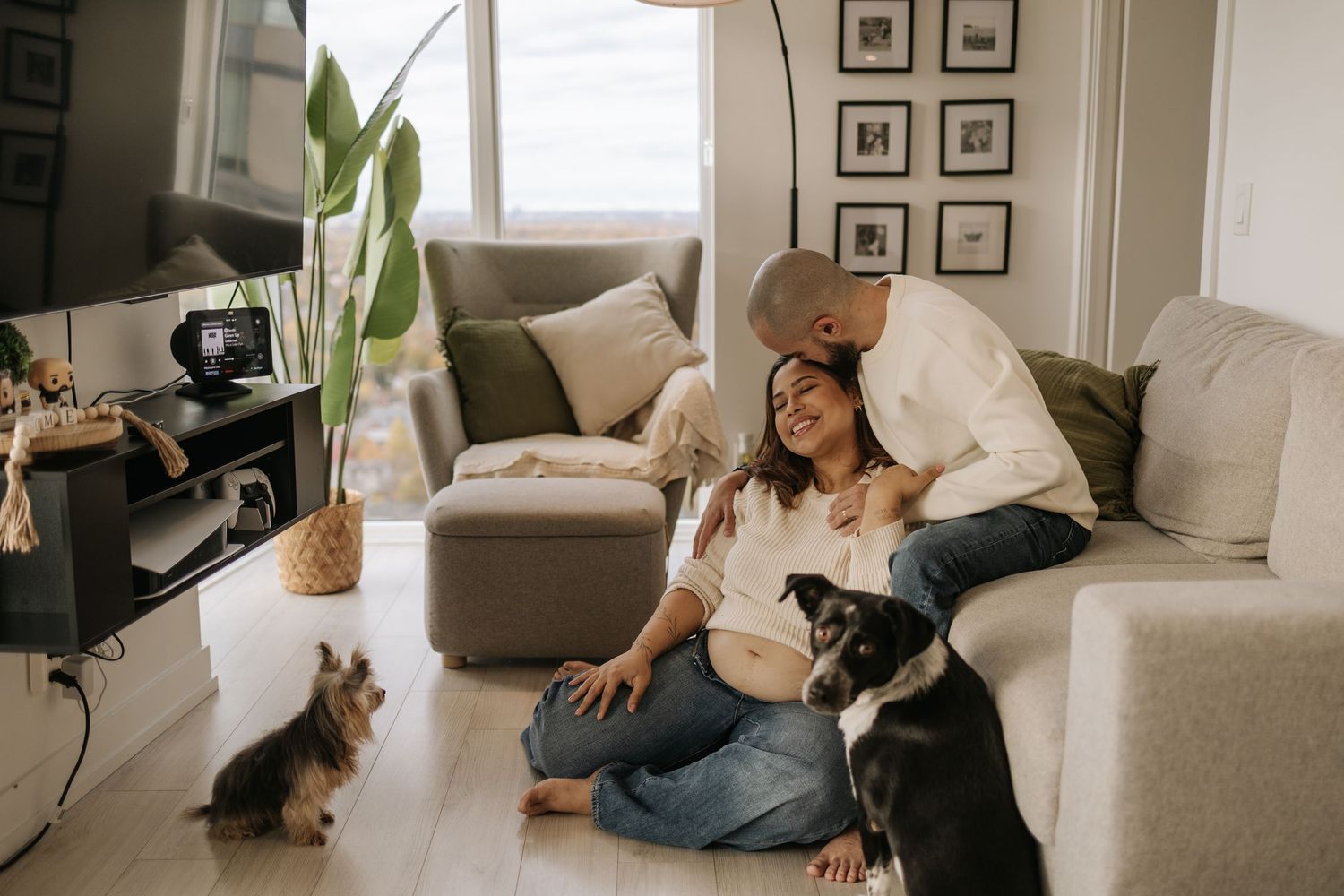 husband on the couch, pregnant with on the floor wearing a white sweater and jeans, showing ther prenant belly, and their 2 dogs with them.