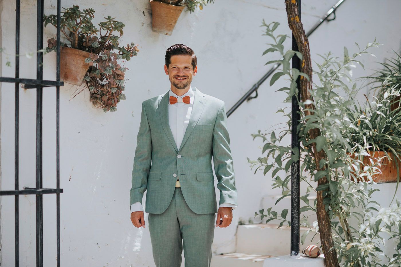 A groom wearing a sage green suit with white bow tie stands smiling in a Mediterranean-style courtyard setting.