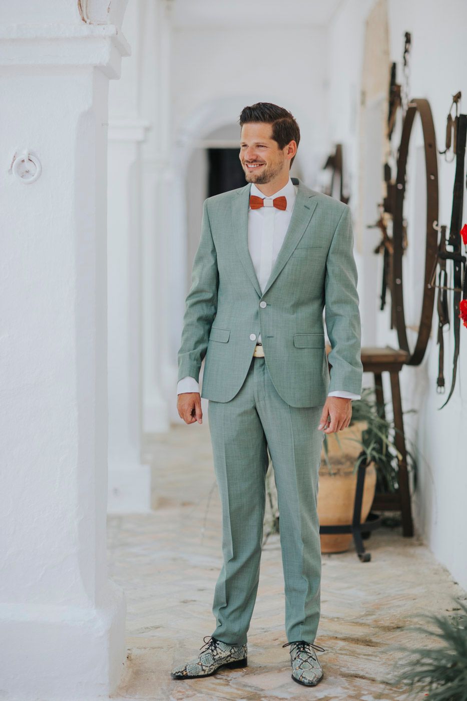 A sage green suited figure poses in a white hallway wearing unique patterned dress shoes and coral bow tie.