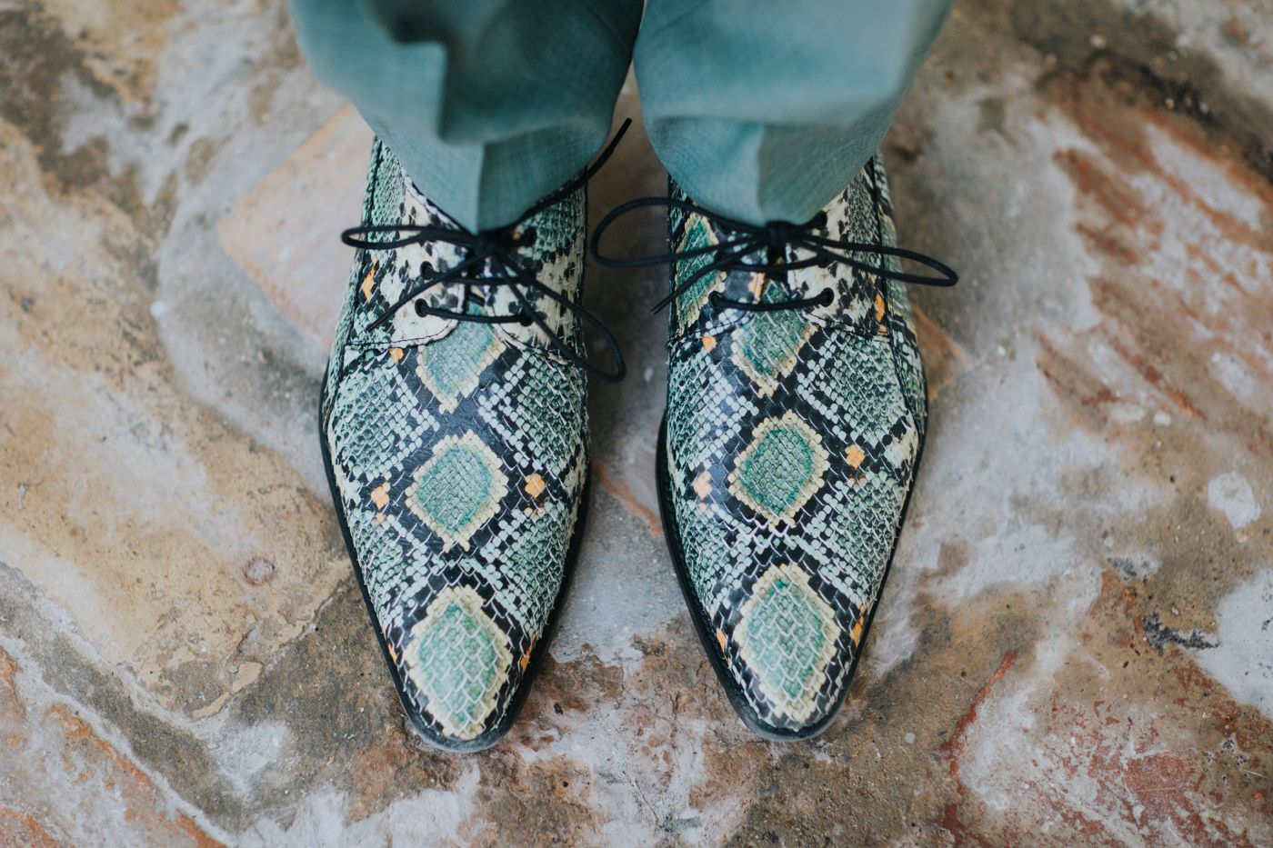 A pair of pointed-toe snakeskin pattern shoes in mint green and black colors photographed on a textured stone surface.