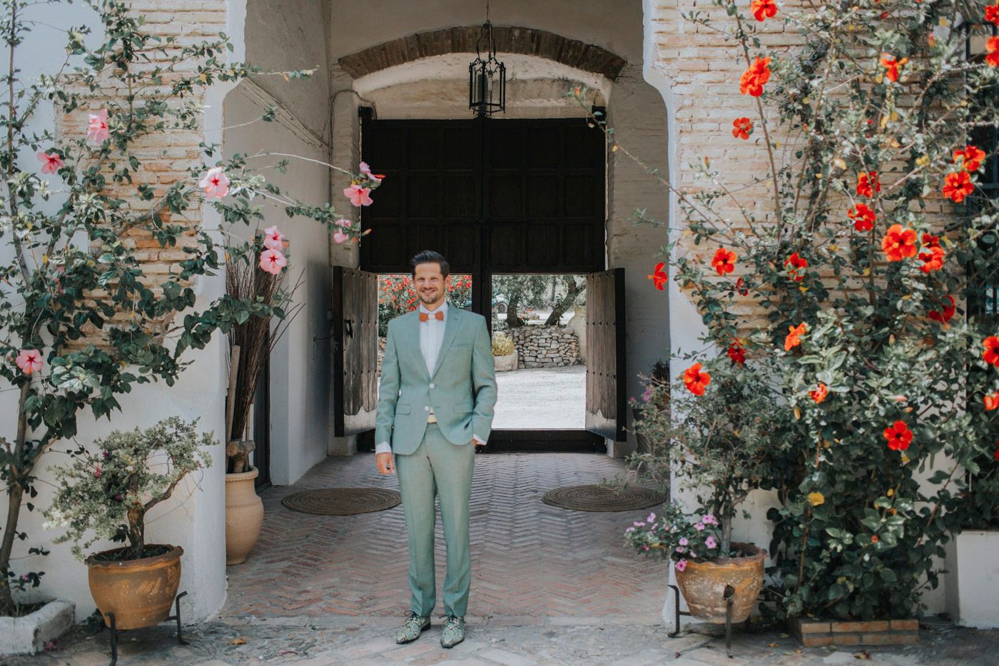 A person in a light blue suit stands in an archway entrance surrounded by blooming red flowers and climbing vines.