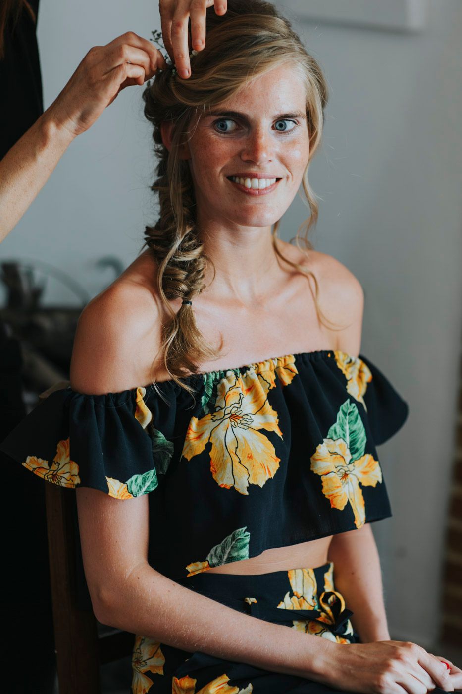 Someone wearing a black off-shoulder crop top with yellow floral print gets their hair styled in a side braid.