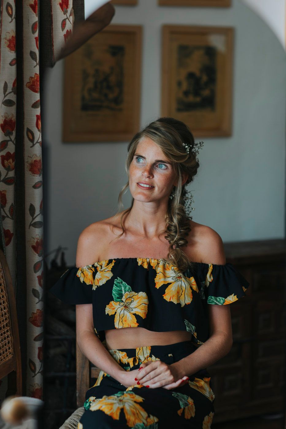 Woman in black and yellow floral off-shoulder dress sits thoughtfully in a room with framed artwork and curtains.