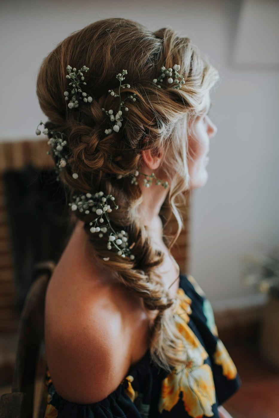 Elegant bridal hairstyle with baby's breath flowers woven through a side-swept braid shown against dark floral dress.