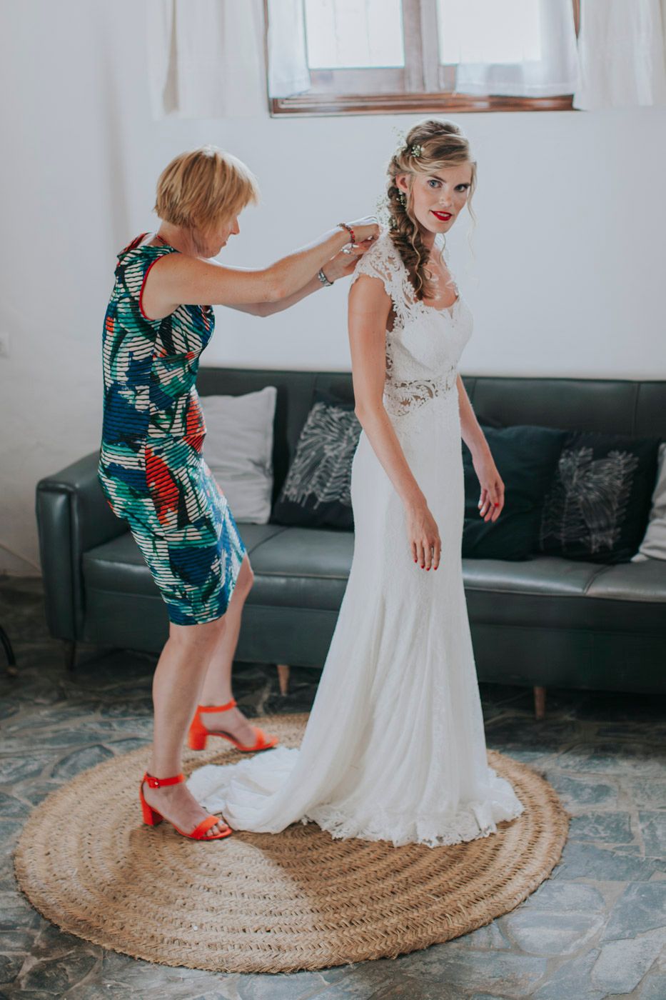 A person in a floral dress helps adjust a stunning white lace wedding gown on a round jute rug beside a gray couch.