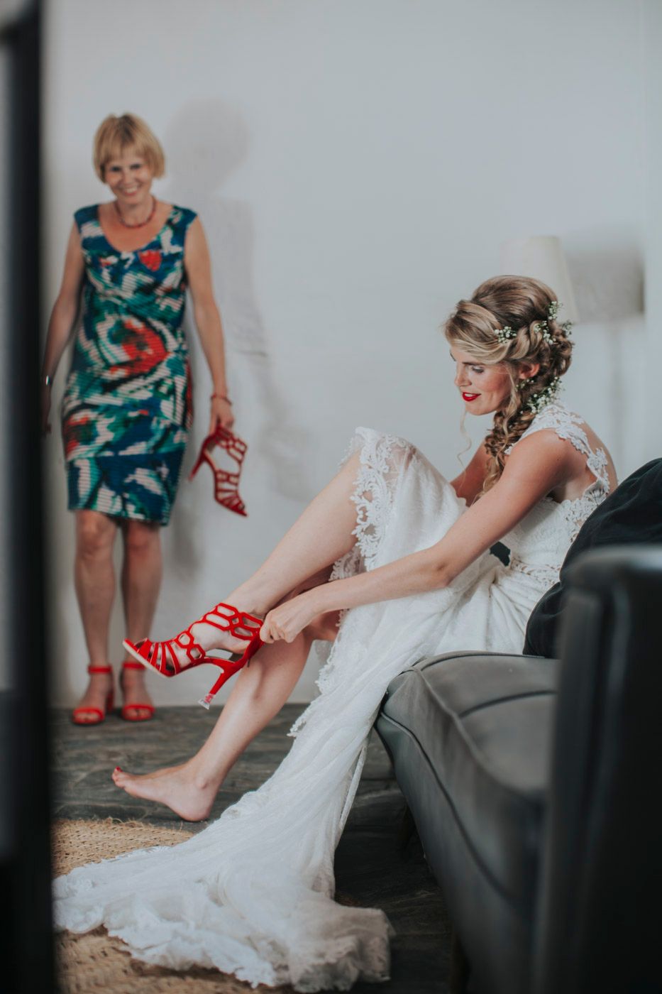 A bride in a lace wedding dress puts on red heels while someone in a floral dress stands nearby with matching shoes.