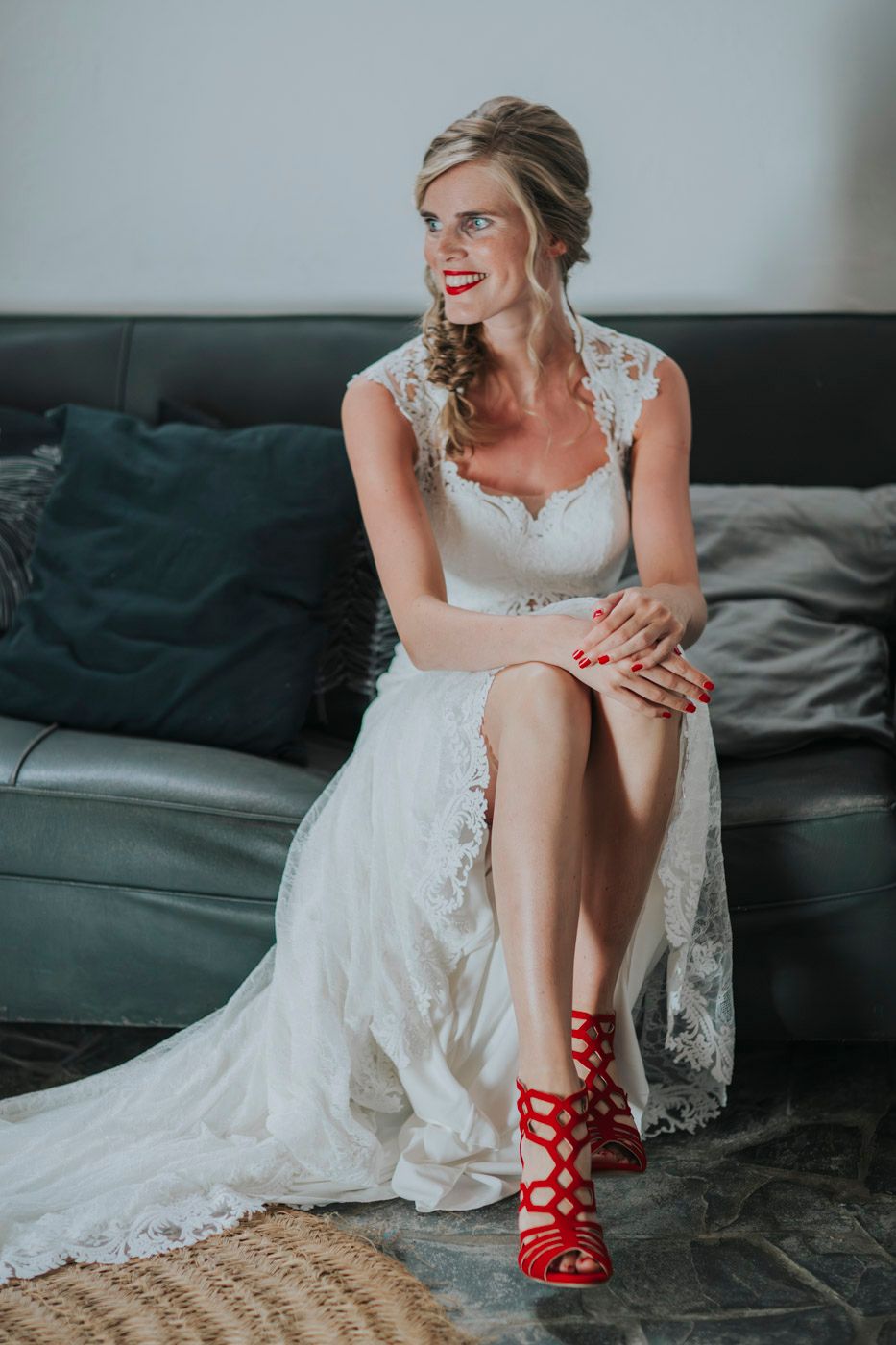 Person in elegant lace wedding dress seated on gray couch wearing vibrant red strappy heels and smiling.