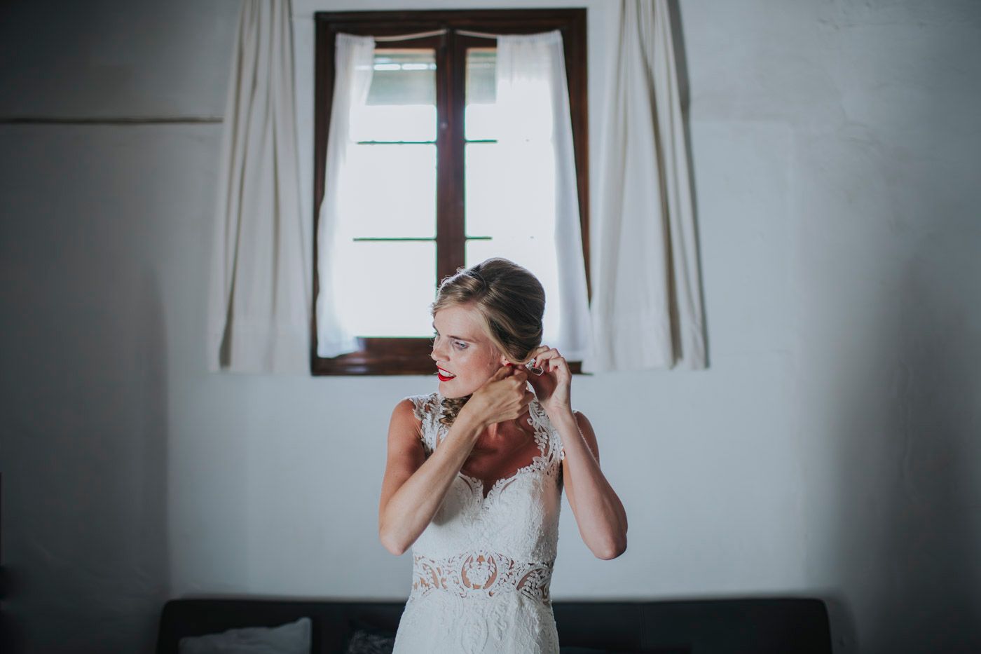 A bride in a white lace wedding dress puts on her earrings near a window in a simple room.