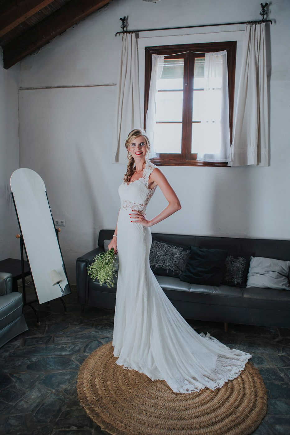 A person in an elegant white wedding dress with a long train poses on a round jute rug in a rustic interior setting.