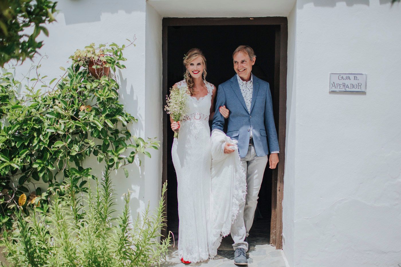 A couple walks out of a doorway of a white building surrounded by greenery and blooming flowers on a sunny day.