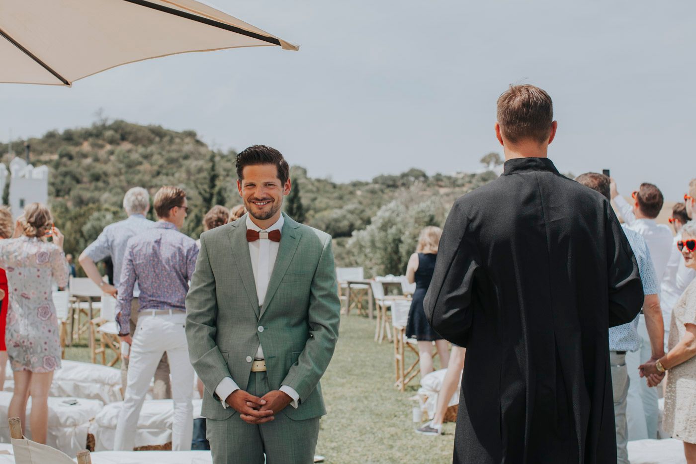 Outdoor wedding scene in a grassy field with guests gathered under umbrellas and a person in a sage green suit.