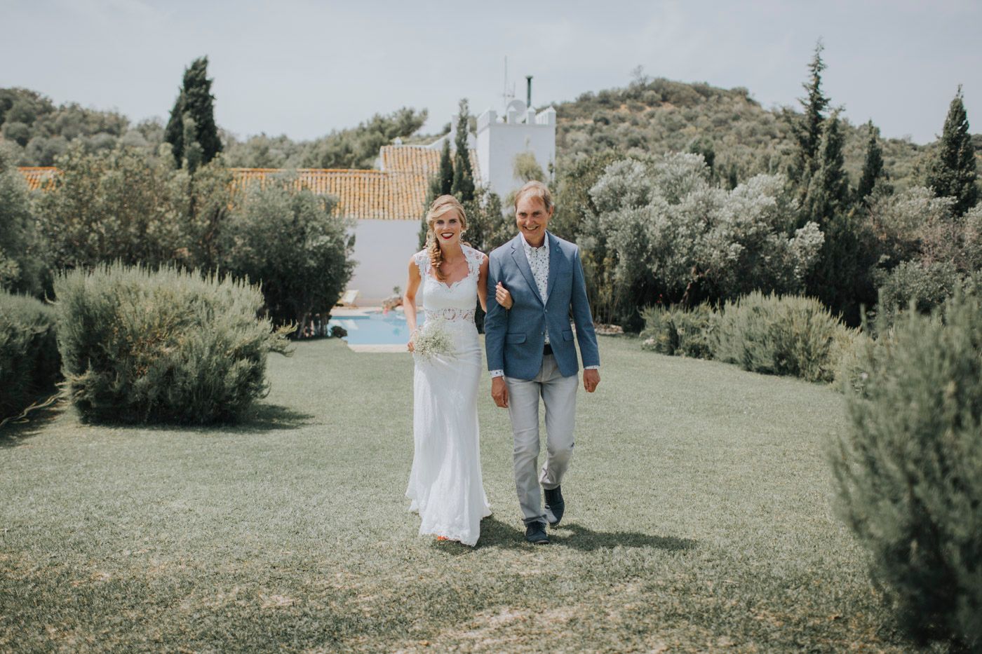 A couple walks hand in hand across a lush garden lawn during an outdoor wedding ceremony.