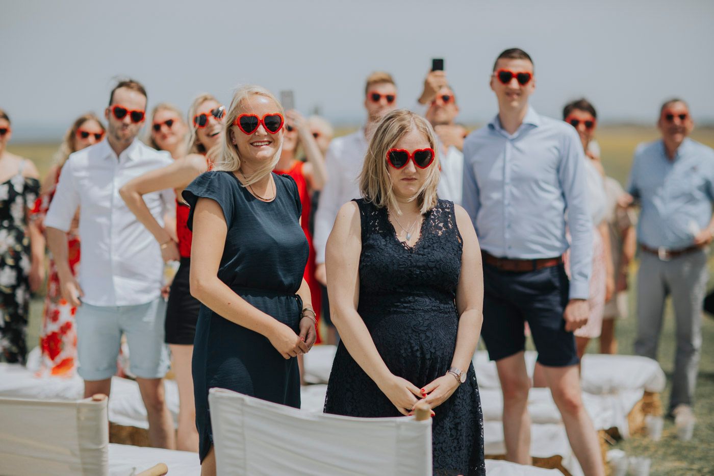 Wedding guests in summer attire gather on a seaside deck wearing sunglasses and navy blue outfits on a bright sunny day.