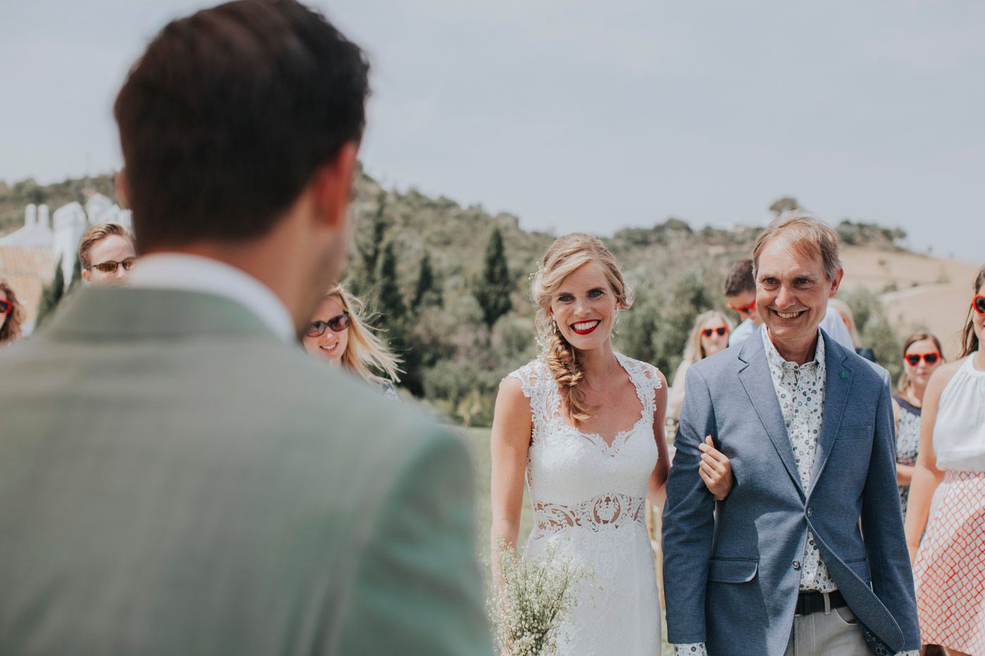 A couple in wedding attire stands outdoors during a sunny ceremony with hills in the background.