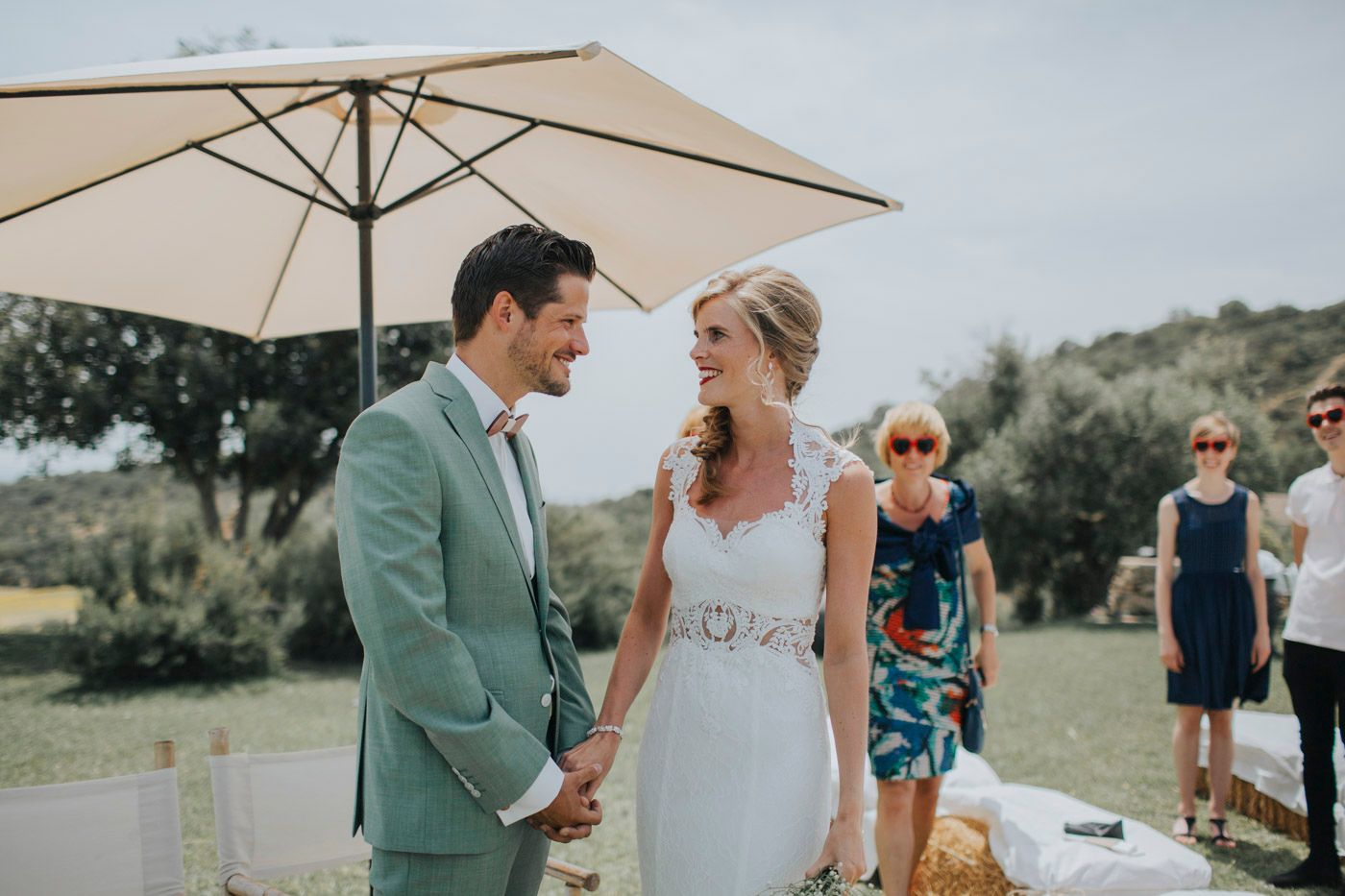 Couple in wedding attire stands under white umbrella at outdoor garden venue on sunny day.